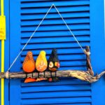 3 passarinhos de tecido amarelo, laranja e preto, pousados num galho de madeira