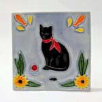 azulejo em relevo de gato preto de lenço vermelho ao pescoço, num fundo azul um ratinho uma bola vermelha e flores