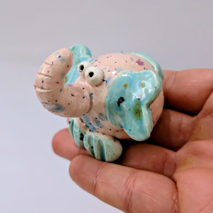 mão a segurar um boneco de elefante em ceramica moldado á mão em cor de rosa e azul